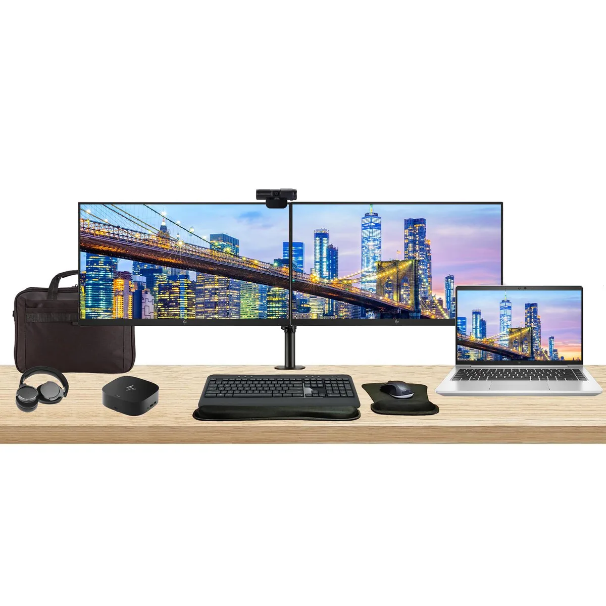 Closed silver laptop beside two black monitors on a shared stand with dock keyboard mouse webcam headphones and briefcase