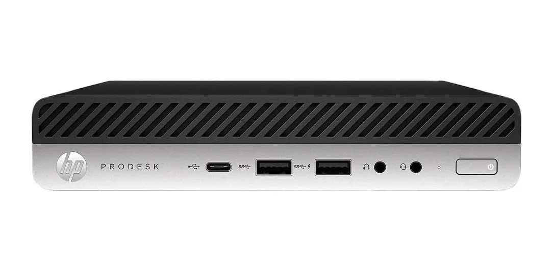 Small silver mini PC box, compact square shape, matte finish, front USB and audio ports, rear DisplayPort and Ethernet visible